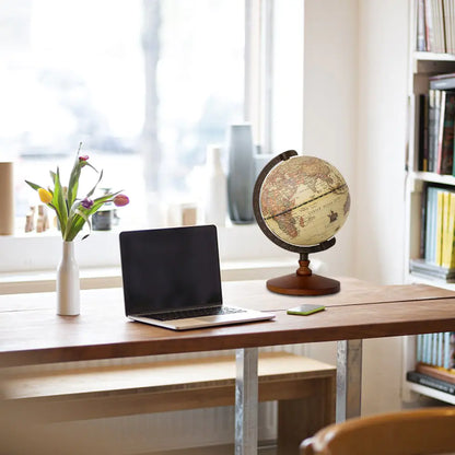 Globe on a desk with a laptop and flowers in a room with a window and bookshelf.